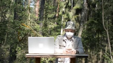 Koruyucu maskeli bir adam ormanda oturur ve telefonla SMS yazar, COVID-19 dünya salgını konsepti.