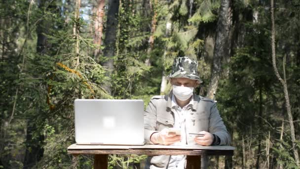 Un homme avec un masque de protection est assis à une table avec un ordinateur portable dans la forêt et paie l'achat avec une carte de crédit, concept de pandémie de coronavirus COVID-19 — Video