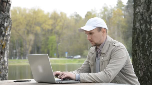 Jonge Kaukasische man werkt aan een laptop over de natuur op de achtergrond van een bosmeer, coronavirus COVID-19 pandemie concept, zelf-isolatie — Stockvideo