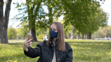Şehir parkında video kaydı için akıllı telefon kullanan koruyucu maskeli genç bir kadın.
