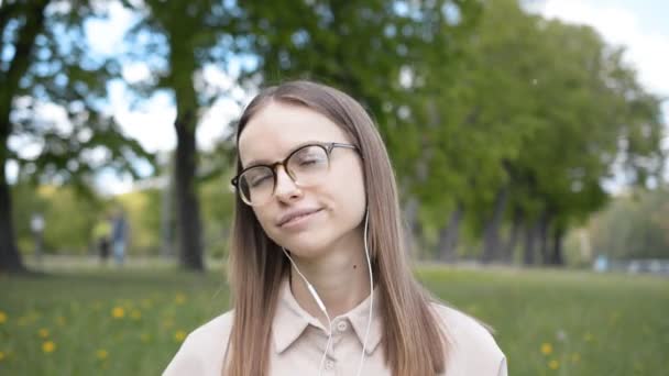 Retrato de una hermosa estudiante en gafas en un parque de la ciudad escucha música favorita — Vídeos de Stock