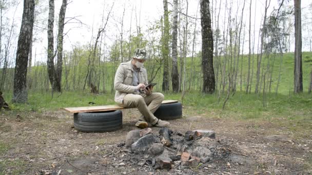 Um homem com uma máscara médica senta-se sozinho na floresta e olha para algo em seu tablet — Vídeo de Stock