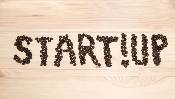 The word - start up - is made of roasted coffee beans on a light wooden textured surface of the table.
