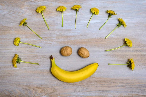 A comic image of a smiling face on a textured wooden surface. Banana is a smile, walnuts are eyes and dandelions are like a voluminous hairstyle.