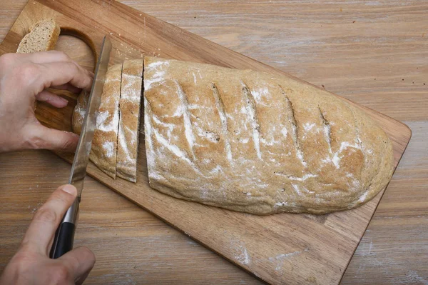 Female hands that neatly hold a large piece of cut gray hand-made fresh bread sprinkled with white flour on a wooden board and continue to cut it into small pieces with a large knife, top view.