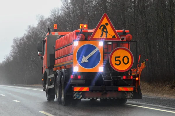 Road work machine truck with lighting direction sign and speed limit 50 on right lane driving on highway road on spring evening