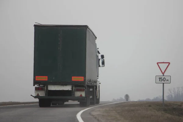 Heavy truck with dark green semi trailer drive on suburban asphalted road in fog at spring day, rear-side view close up  international logistics, cargo transportation, trucking industry