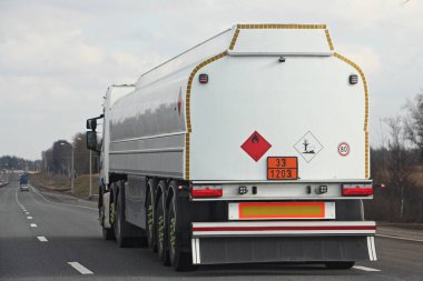 33 / 1203 tehlikeli sınıf işaretli beyaz tır yakıt tankeri ve asfalt yolda boş bir varil sürücüsü mavi gökyüzü arka planında, yan görüş ADR tehlikeli kargo