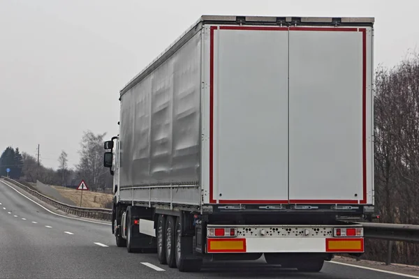 Modern European white semi truck with copy space blank on three-axle trailer rear door on empty highway road at spring day