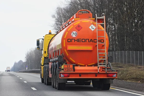 Modern bright orange semi truck fuel tanker with 33 1203 dangerous class sign and Russian inscription INFLAMMABLE drive on suburban empty road at spring day, rear view - ADR hazardous cargo in Russia