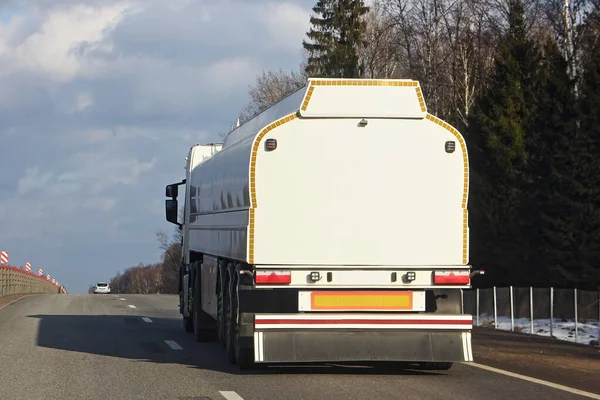 Modern white fuel truck gasoline tanker moving on asphalt highway on a spring day, rear side view - ADR dangerous cargo, copy space for text on barrel back surface