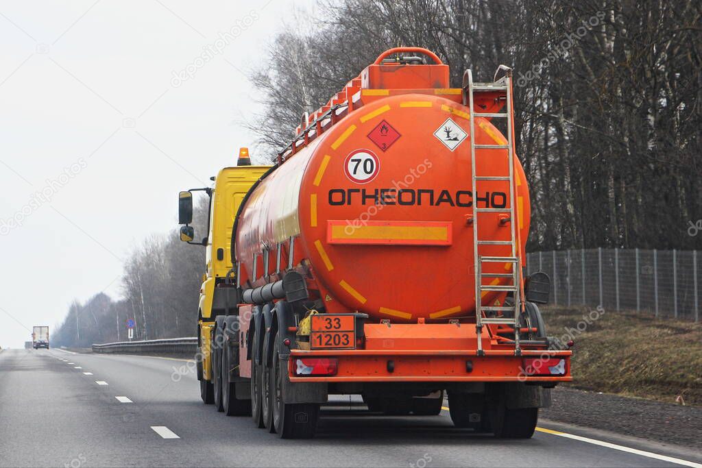 Modern bright orange semi truck fuel tanker with 33 1203 dangerous class sign and Russian inscription INFLAMMABLE drive on suburban empty road at spring day, rear view - ADR hazardous cargo in Russia