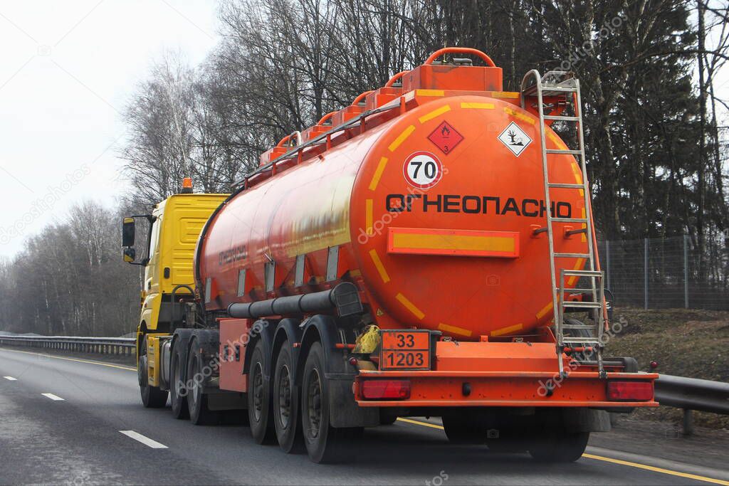 Modern bright orange semi truck fuel tanker with 33 1203 dangerous class sign and Russian inscription FLAMMABLE drive on highway at spring day in perspective, rear side view - ADR hazardous cargo