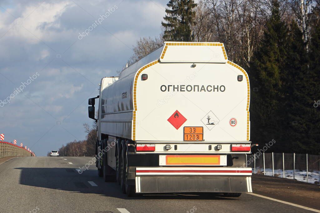 White semi truck fuel tanker with 33 1203 dangerous class sign and inscription in Russian FLAMMABLE moving on suburban asphalt highway road on a spring day, side rear view - ADR liquid hazardous cargo