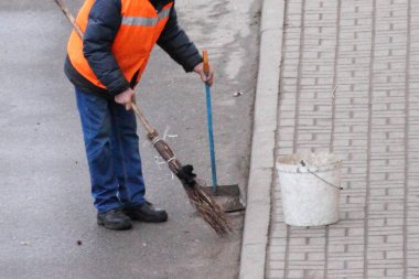 Turuncu yelekli bir hademe kaldırımlı sokağı süpürgeyle süpürüyor.