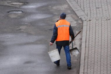 Turuncu yelekli bir hademe elinde kovayla yürür ve asfalt cadde boyunca kepçeyle dolaşır.