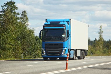 Beyaz römorklu mavi kabin kamyonu, banliyö otobanının ön tarafı manzaralı yaz günü ağaçlar ve mavi gökyüzü arka planı