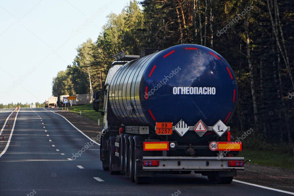 Dark blue semi truck bitumen tanker with 99/3575 dangerous class sign and Russian inscription FLAMMABLE on barrel drive on asphalt highway on a summer day on forest background, ADR hazardous cargo