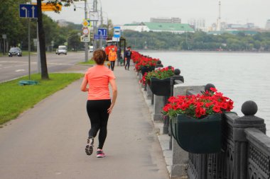 Açık hava eğitim aktivitesi, şeftali rengi spor giysili bir kadın yaz akşamı Moskova nehrinin yanındaki toprak toprak yolda çit boyunca koşuyor.