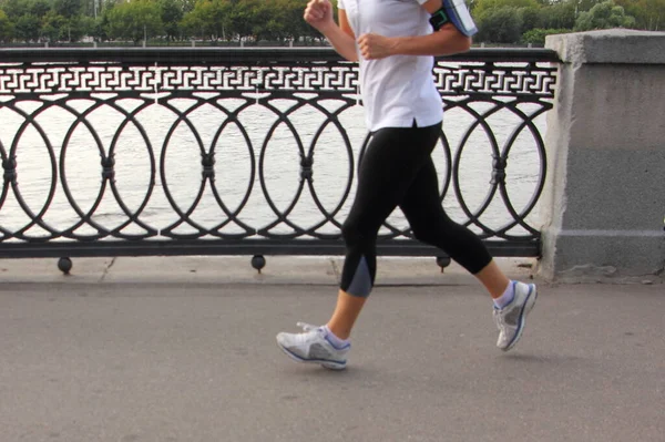 Running man in black-white sportswear and sportshoes jogging along the fence of embankment alley of Moscow river on a summer evening, without a head side view close up, urban activity lifestyle