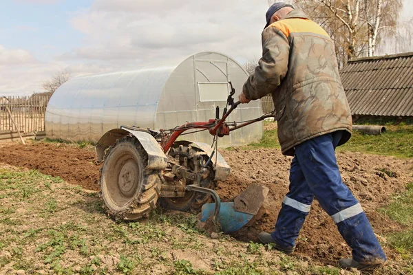 高齢のヨーロッパの男性農家は 温室や古い納屋の背景に春の日に一時停止した耕運機でベッドを耕します — ストック写真