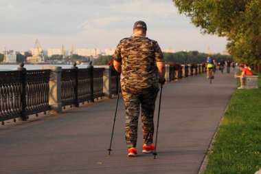 İskandinav yürüyüşü, kamuflajlı bir kadın kayak direkli bir kadın yaz günü Park ara sokaklarında yürüyor, ağaçların arkasına bakıyor ve çitlerin arkasına saklanıyor, açık hava aktiviteleri yapıyor.