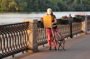 Genç kız sokak ressamı, güneşli bir yaz akşamında, boş bir ara sokakta, rıhtımdaki parkta resim çiziyor. Amatör sanat eseri.