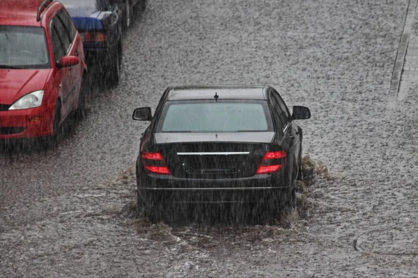 黒い車は駐車車両の背景に街の大雨の間に庭の大きな水たまりを通って運転しています 後部トップビュー — ストック写真