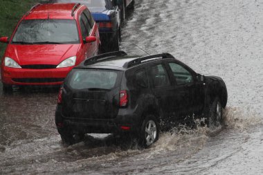 Siyah bir SUV arabası, bir bahar günü, şehrin arka tarafındaki bir sokakta sağanak yağmurdan sonra park halindeki arabaların yanındaki derin bir su birikintisinde sürüyor.