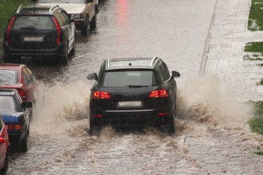 Koyu renk bir SUV arabası, şehrin bahar günü, yoğun yağmurda park halindeki araçların yanındaki büyük su birikintisine su sıçratarak hızlı gider.