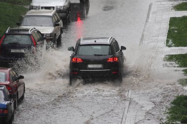 Siyah bir SUV arabası, yoğun bir su birikintisinde, şehrin bir bahar gününde, bir sokakta, park halindeki arabaların yanındaki derin bir su birikintisinde hızla araba sürüyor.