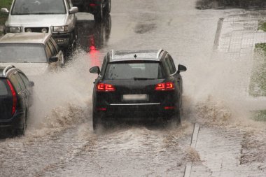 Siyah bir Alman lüks SUV aracı, bir bahar günü, arka planda sağanak yağmurda, park halindeki arabaların yanında, sokağın aşağısına doğru ağır bir sprey püskürtüyle hızla gider.
