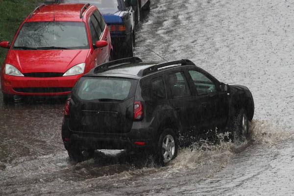 黒いSuv車が春の日に街の路上で激しい雨の後 駐車車の横に深い水たまりの中を運転しています リア側のビュー — ストック写真