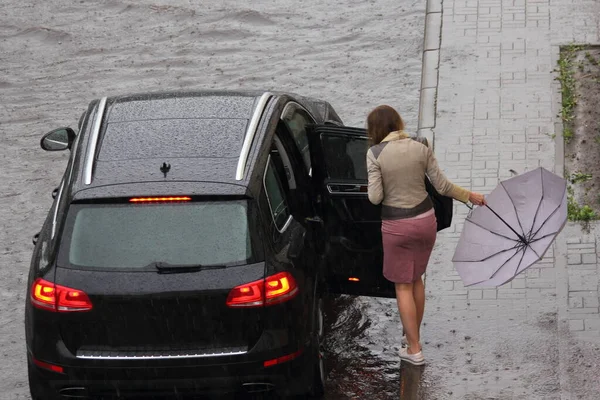 A young Caucasian woman with pink umbrella gets into right rear door a luxury SUV black car from the sidewalk on a road in the city on a spring day in a deep puddle after rain