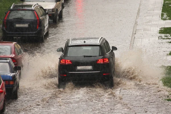 暗いSuv車は 春の日に街の大雨の中で駐車車両の横にある通りの下の大きな水たまりにスプラッシュ水で高速ドライブ 都市オフロードバックビュー — ストック写真