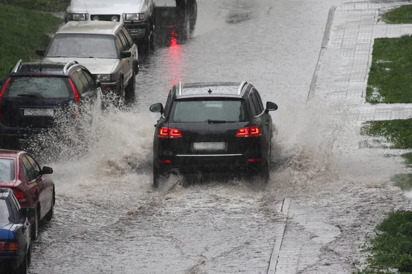 黒いSuv車は 春の日に街の路上で大雨の中で駐車車の横に深い水たまりに重いスプレーで高速運転しています リアビュー — ストック写真