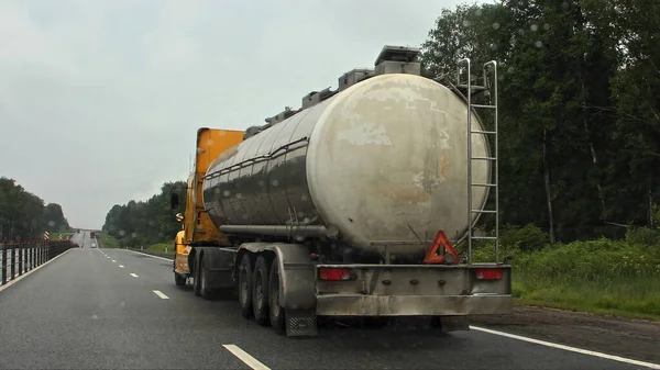 Gasoline and oil transportation, old chrome gray barrel fuel tank semi trailer with American truck on suburban highway road at summer day on green forest on roadside background, rear side view closeup