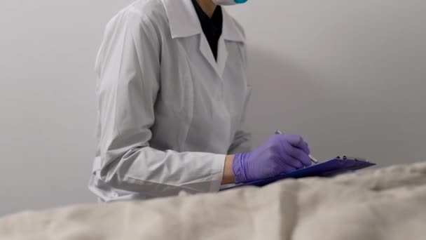 Άρρωστη γυναίκα είναι ξαπλωμένη στο κρεβάτι στο δωμάτιο του νοσοκομείου, βήχοντας με μάσκα στο πρόσωπο, ο γιατρός συμπληρώνει το ερωτηματολόγιο — Αρχείο Βίντεο