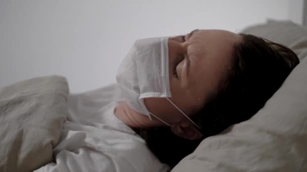 Sick woman with medical mask is coughing hardly lying in bed in hospital room, closeup of face — Stock Video