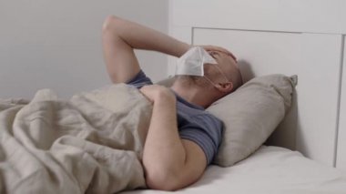 Grip olmuş erkek hasta yüzüne maske takmış. Yatakta uzanıyor ve hastane odasında öksürüyor.