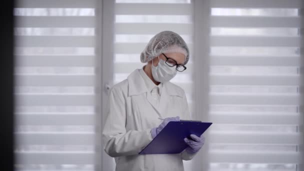 Covid-19 pandémico. Retrato de un médico con una máscara protectora sobre el fondo de una ventana blanca . — Vídeo de stock