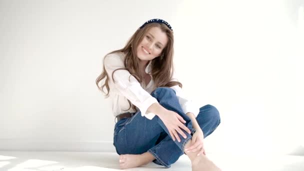 Joven mujer sonriente tímida atractiva con el pelo rizado largo se sienta en un piso ligero contra un fondo claro en una chaqueta blanca y jeans — Vídeos de Stock