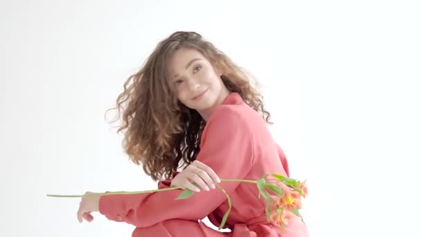 Retrato de una joven sonriente con el pelo largo y castaño en un traje rojo brillante, sostiene una flor naranja brillante sobre un fondo brillante en la habitación — Vídeos de Stock