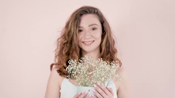 Retrato. Hermosa joven con el pelo castaño rizado sobre un fondo claro con flores en las manos y sonriendo — Vídeos de Stock