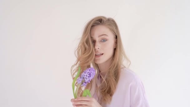 Retrato de una joven atractiva mujer rubia delicada con el pelo largo y rizado sobre un fondo claro, que muestra emociones, sonrisas, sostiene una flor — Vídeos de Stock