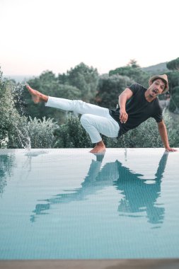 Siyah gömlekli, şapkalı, yüzme havuzunun yanında suyla oynayan etnik tarzı genç bir adam.