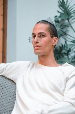 Ekzotik, beyaz, yuvarlak, şeffaf güneş gözlüklü genç bir adam, tropik bir bitkinin önündeki koltukta otururken kameraya bakıyor.