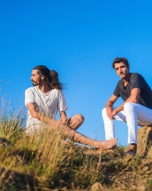 Farklı yönlere bakan etnik ve beyaz gençler arka planda açık mavi bir gökyüzü ile doğada oturuyorlar.