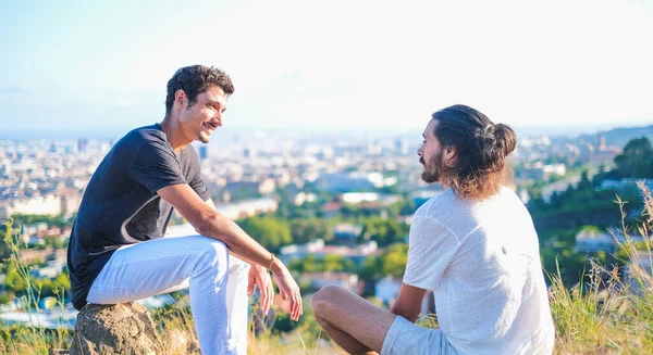 Ethnic and caucasian young mans looking each other in a profound way having a good conversation while sitting in the nature with a city on the background