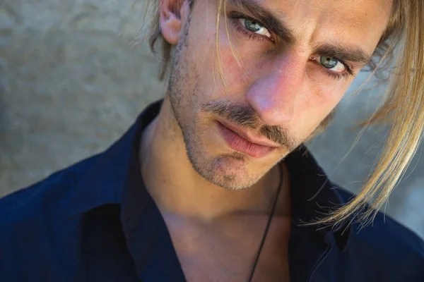 Young male model with medium long blonde hair, wearing a necklace and blue eyes, dressed in a shirt and looking directly at the camera.